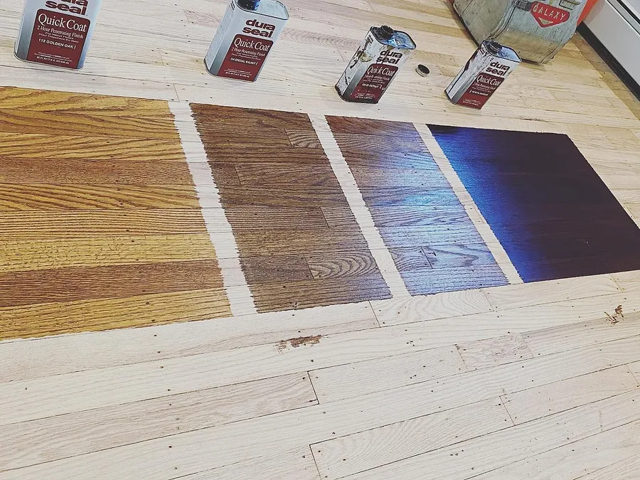 Four stain color samples on a freshly sanded hardwood floor, with the stain cans behind